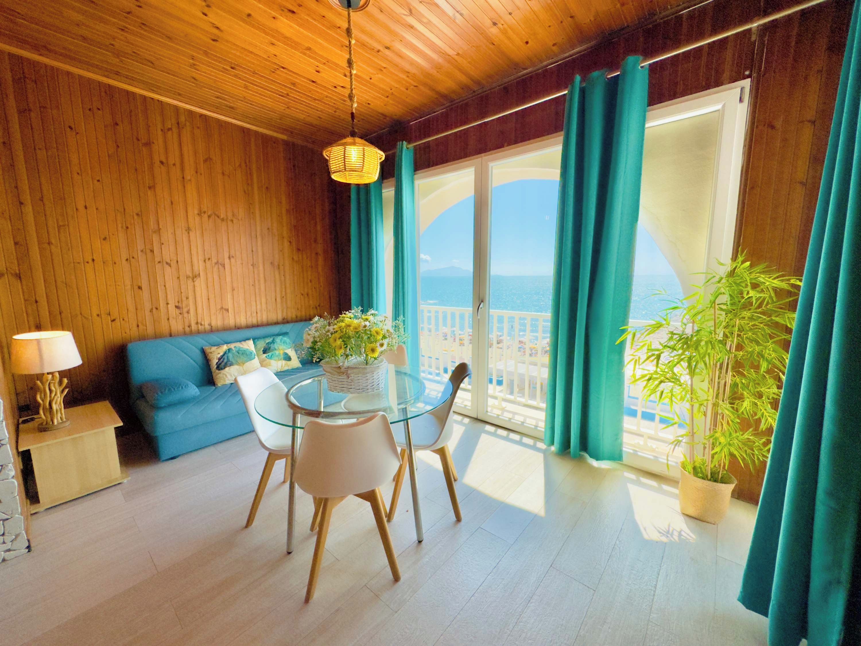 Ein Zimmer mit Holzboden, einem runden Glastisch mit Stühlen, einer Couch mit Kissen, einer Lampe und Blick auf das Meer durch große Fenster.