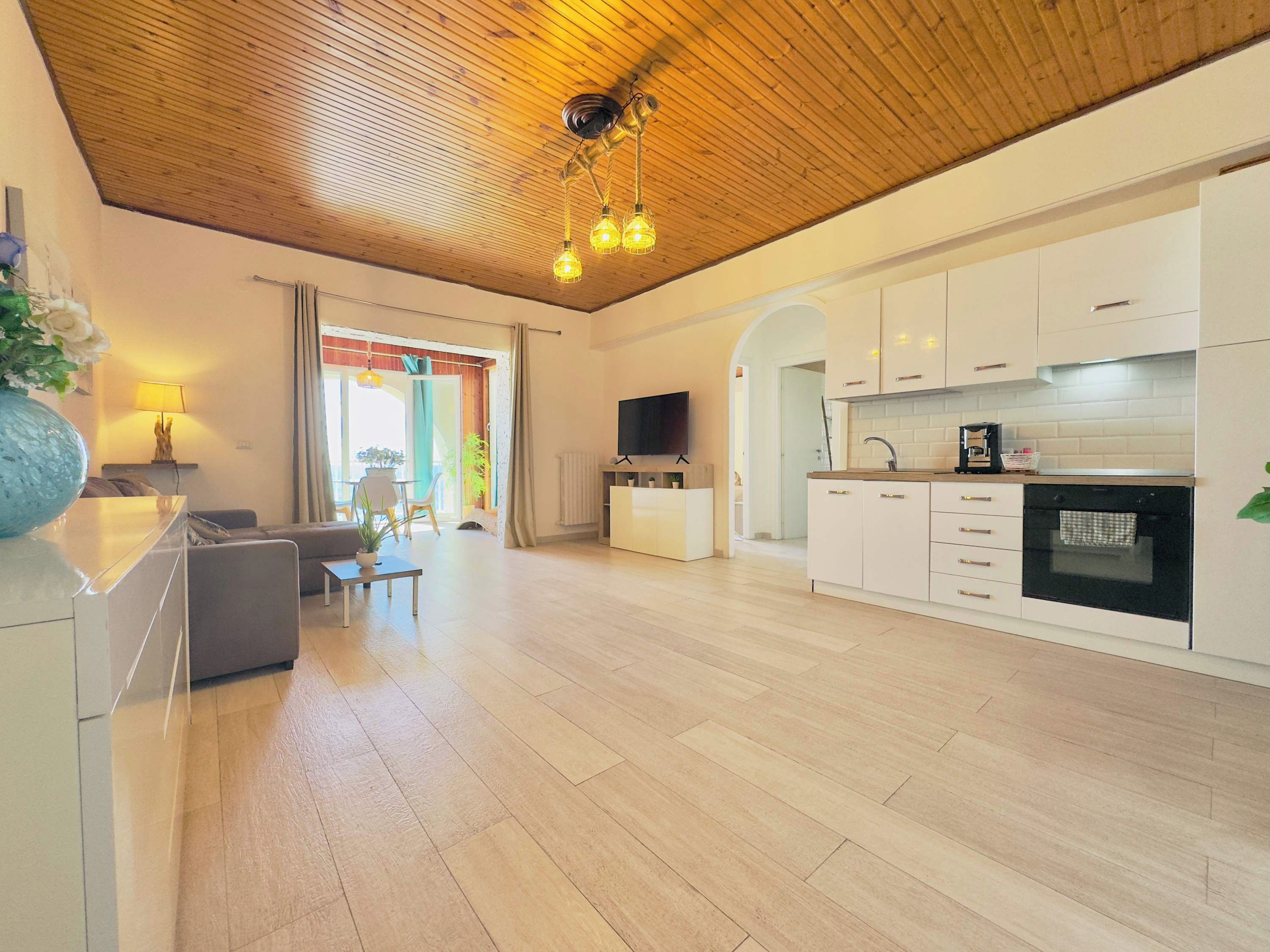 A modern living space with a wooden ceiling, a kitchen with white cabinets, a dining area with a table and chairs, and a TV mounted on the wall.