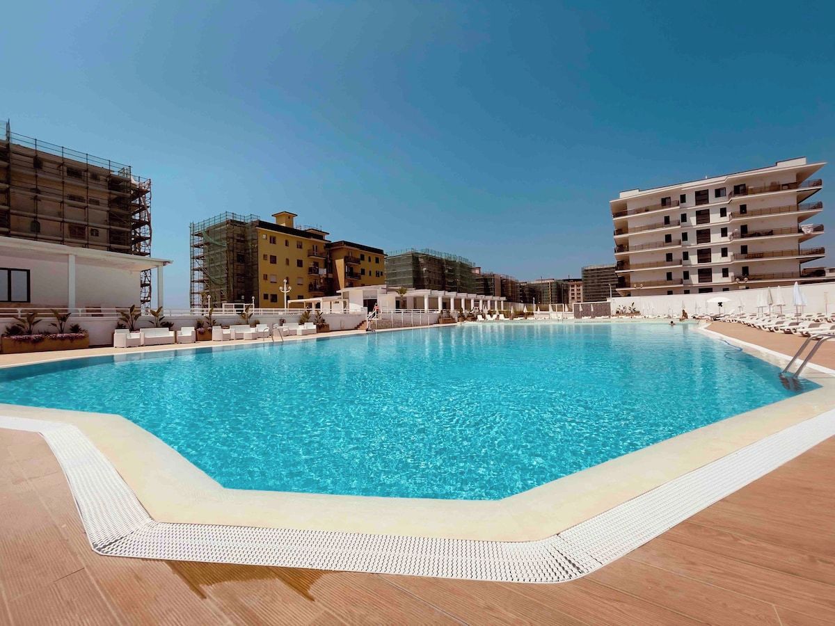 Une piscine dotée d’une terrasse carrelée, entourée d’immeubles avec des balcons, dont certains sont en construction.