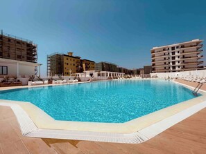 Une piscine dotée d’une terrasse carrelée, entourée d’immeubles avec des balcons, dont certains sont en construction.