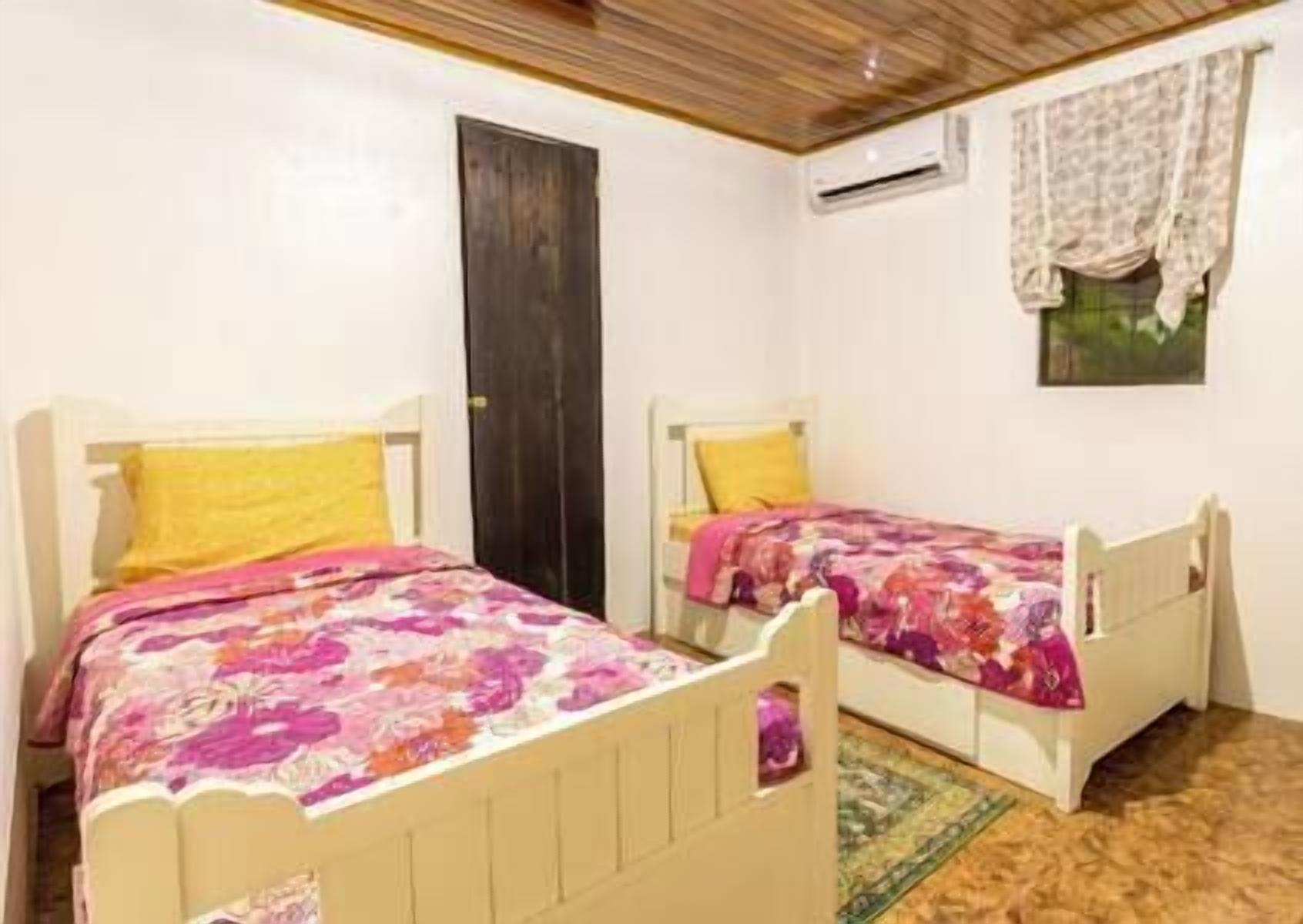 A room with two single beds, each with a floral bedspread and a yellow pillow. There is a wooden door and an air conditioning unit on the wall.
