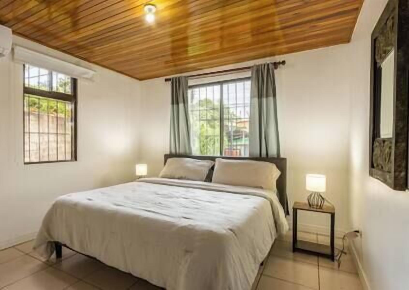 A bedroom with a wooden ceiling, a bed with a white bedspread, two bedside lamps, and a window with curtains.