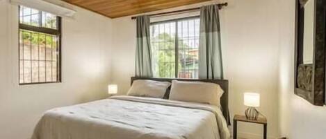 A bedroom with a wooden ceiling, a bed with a white bedspread, two bedside lamps, and a window with curtains.