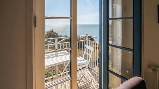 Ein Zimmer mit großem Fenster und Blick auf eine Terrasse mit weißer Brüstung und einer Bank.