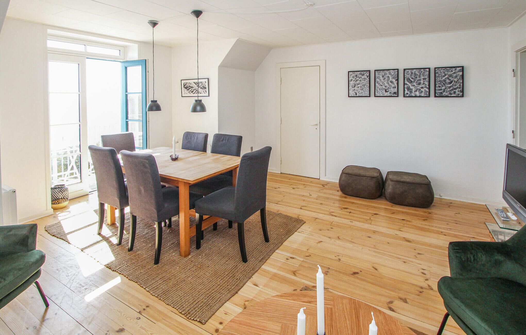 Un coin repas avec une table en bois, six chaises et deux lampes suspendues. On y trouve également quatre œuvres d’art encadrées accrochées au mur, un téléviseur et un canapé.