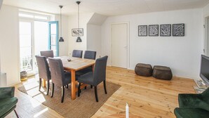 Ein Essbereich mit einem Holztisch, sechs Stühlen und zwei Pendelleuchten. An der Wand hängen vier gerahmte Kunstwerke, ein Fernseher und ein Sofa sind ebenfalls vorhanden.