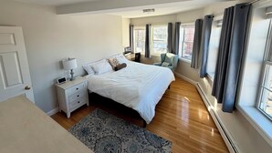 Una camera da letto con un letto, comodini, una lampada, una sedia, una finestra con tende e un termosifone.