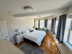 A bedroom with a bed, bedside tables, a lamp, a chair, a window with curtains, and a radiator.