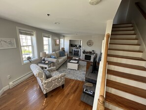 A spacious living area with hardwood flooring, a sofa, a patterned armchair, and a staircase.