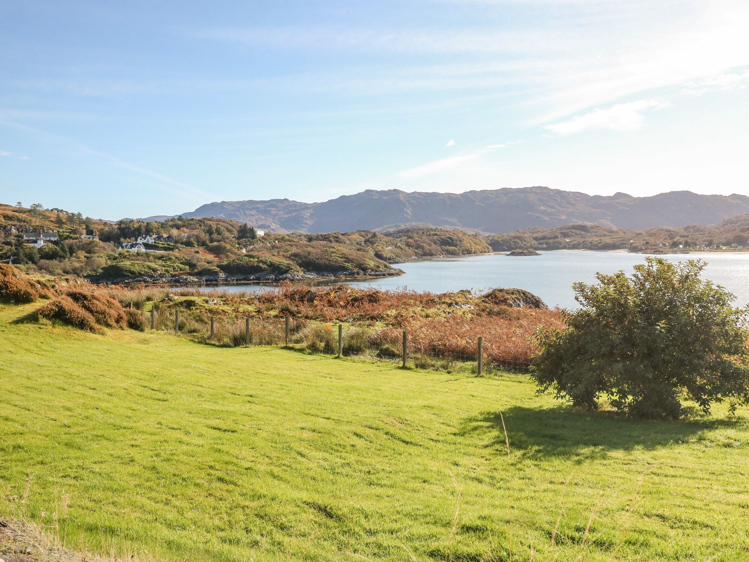 A grassy field with a fence and a tree, leading to a body of water and hills in the background.