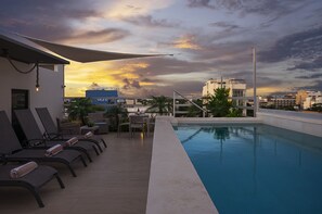 Piscina no terraço com espreguiçadeiras e uma mesa, cercada por edifícios e um céu ao pôr do sol.