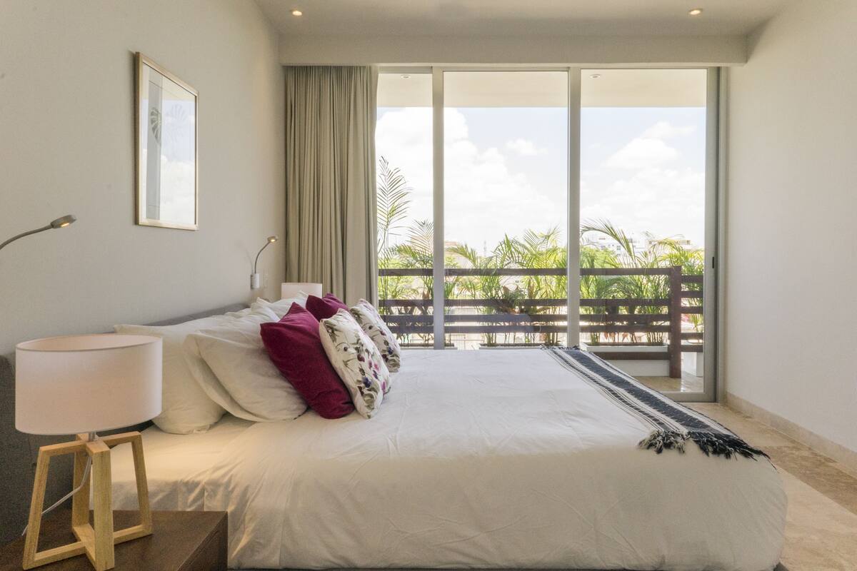 Ein Hotelzimmer mit einem großen Bett, einem Nachttisch mit Lampe und Blick auf einen Balkon mit Palmen.