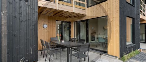 A modern two-story building with a balcony, a dining area with a table and chairs, and large windows.