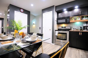 Een moderne keuken met een inbouwoven, magnetron en koffiezetapparaat. Er is een eethoek met een glazen tafel en stoelen, en een plant op de tafel.