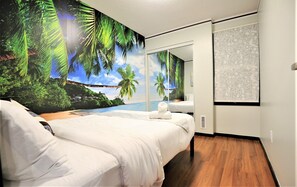 Een slaapkamer met een groot muurschildering van een tropisch strand, een bed, een klein nachtkastje en een raam met een patroon gordijn.