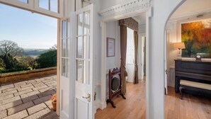 A room with a large open door leading to a balcony with a view of the countryside. There is a piano and a painting on the wall. - Cornish Castle - Luxury Cottages (Saltash)