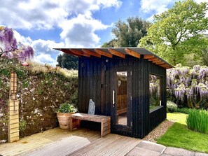 A wooden outdoor structure with a bench and a table, surrounded by a garden with flowering plants and trees. - Cornish Castle - Luxury Cottages (Saltash)
