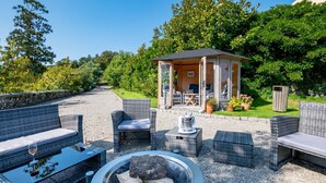 A covered outdoor seating area with wicker furniture, a fire pit, and a small wooden structure. - Cornish Castle - Luxury Cottages (Saltash)