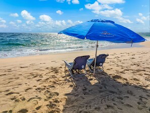 A beach with two beach chairs and a blue umbrella.