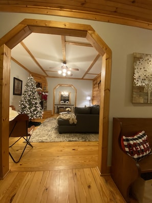 A cozy living room with a wooden ceiling, a fireplace, a Christmas tree, and a dining area in the background.