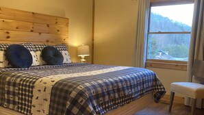 Un dormitorio con cabecera de madera, cama con colcha a cuadros, mesita de noche con lámpara, ventana con vistas a árboles y ventilador de techo.