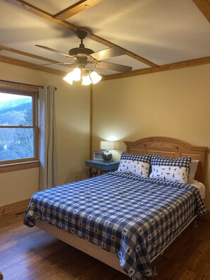 A bedroom with a bed, a ceiling fan, a nightstand, a lamp, and a window with a view of trees.