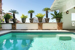 Una piscina amb aigua cristal·lina i blava, envoltada per una paret de maó i coronada amb plantes grans en testos.