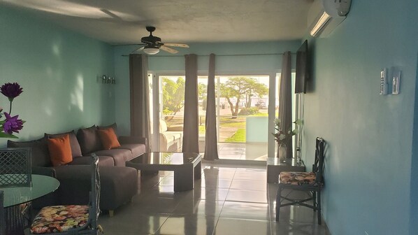 A living room with a sofa, a coffee table, a TV, and a chair. There is a window with curtains, and a ceiling fan is visible. - NEWLY RENOVATED Ocean Front 2 bedroom/ 2 bath, pristine water, divers paradise. (Kralendijk)