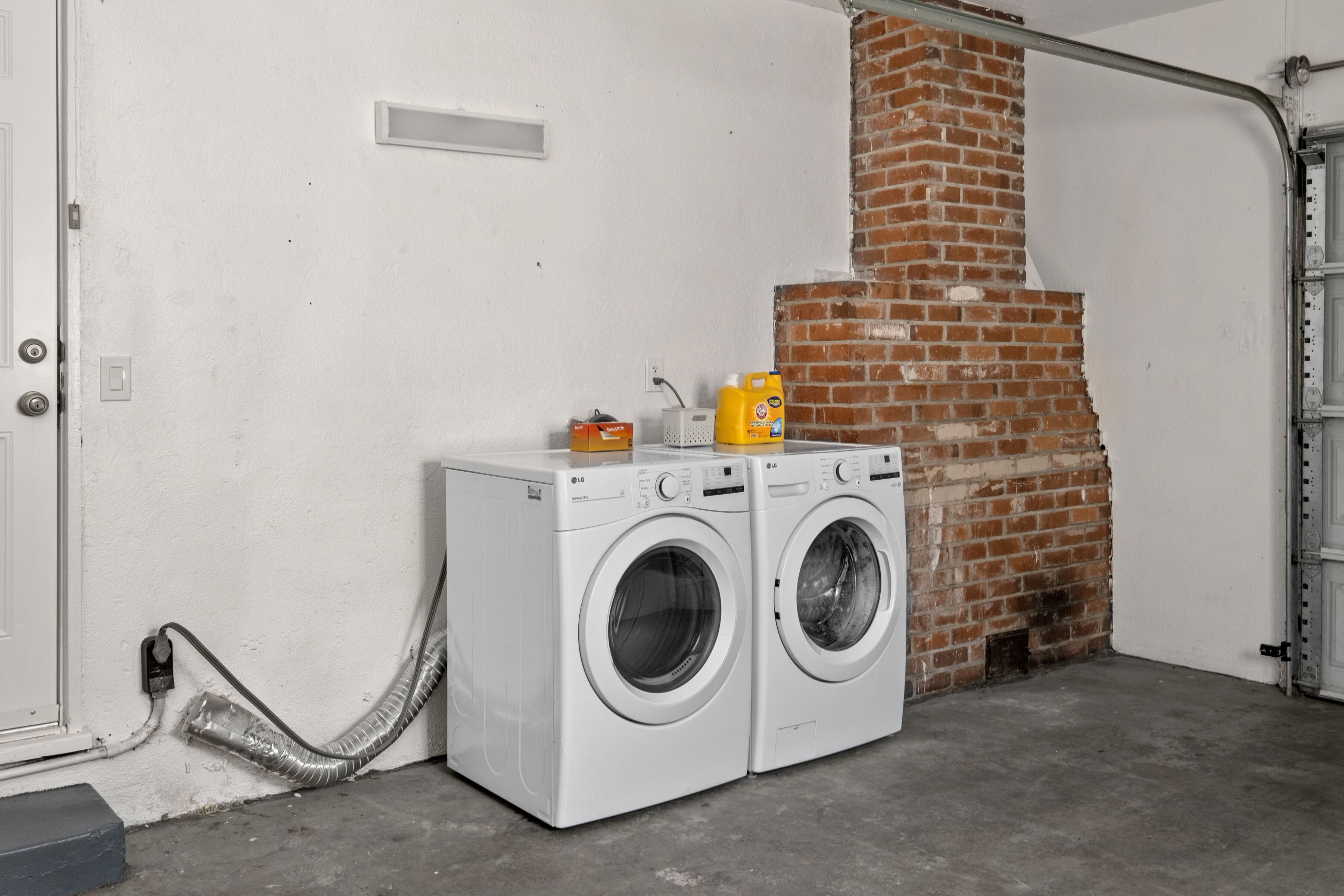 A laundry area with two white front-loading washing machines, a brick wall, and a metal garage door.
