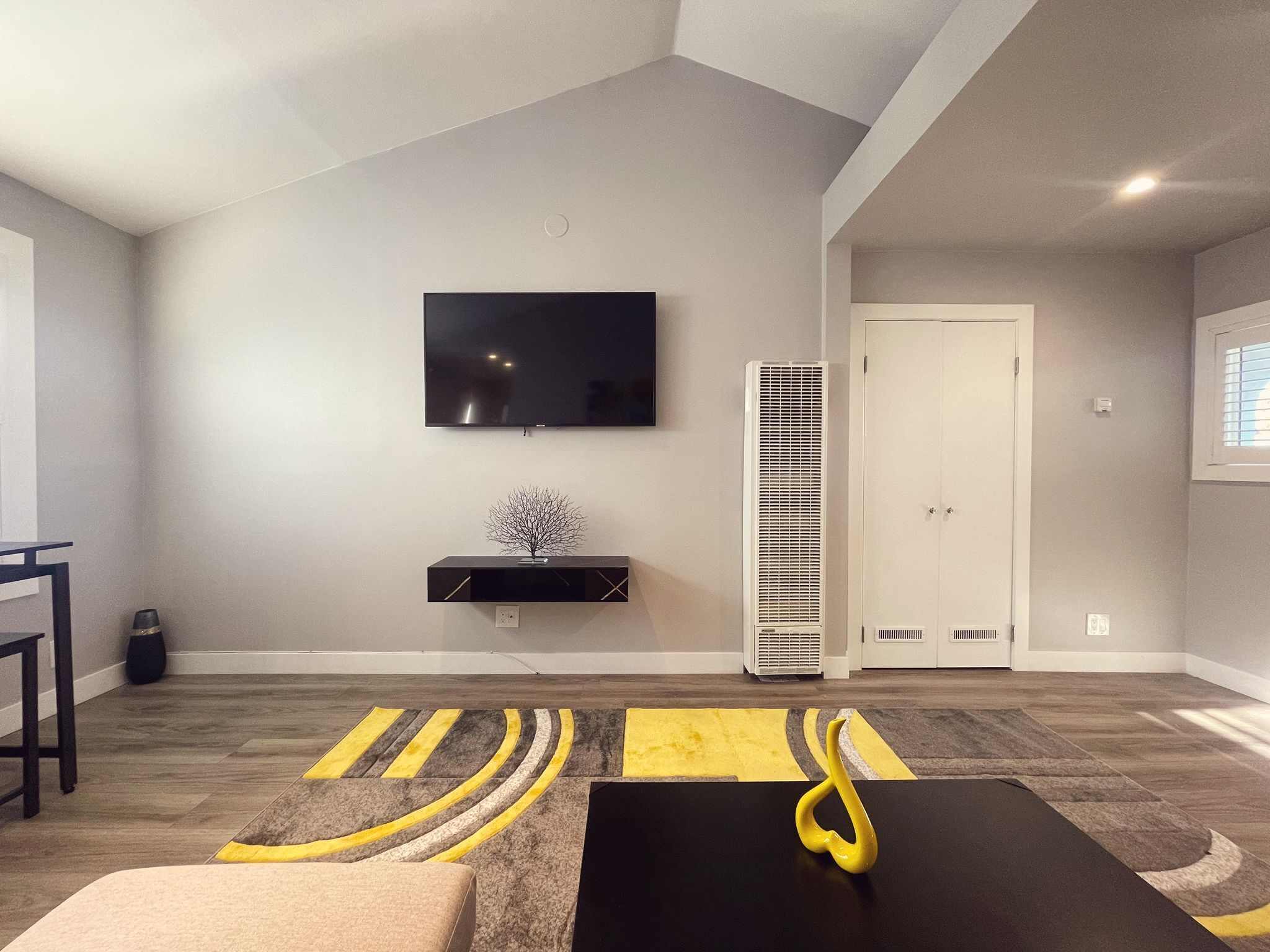 A modern living room with a flat-screen TV mounted on the wall, a black entertainment unit below, a tall white air conditioner, and a patterned rug.