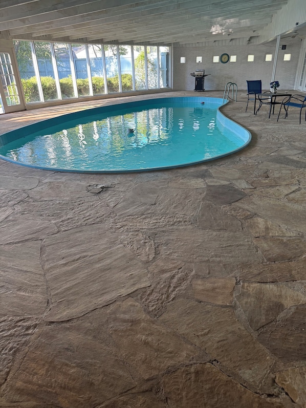 An indoor swimming pool with a stone floor, large windows, and a grill on the wall.