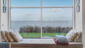 Ein Zimmer mit einem großen Fenster, einer gepolsterten Sitzbank mit geometrisch gemusterten Kissen und Blick auf eine Wiese und Wasser.