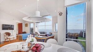Ein modernes Wohnzimmer mit großem Fenster, einem Essbereich mit weißem Tisch und einer gemütlichen Sitzecke mit blauem Sofa und Holzstühlen.