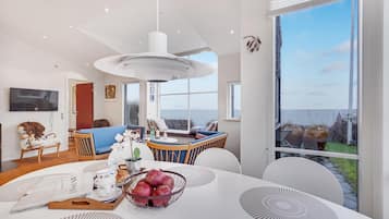 A modern living room with a large window, a dining area with a white table, and a cozy seating area with a blue sofa and wooden chairs.