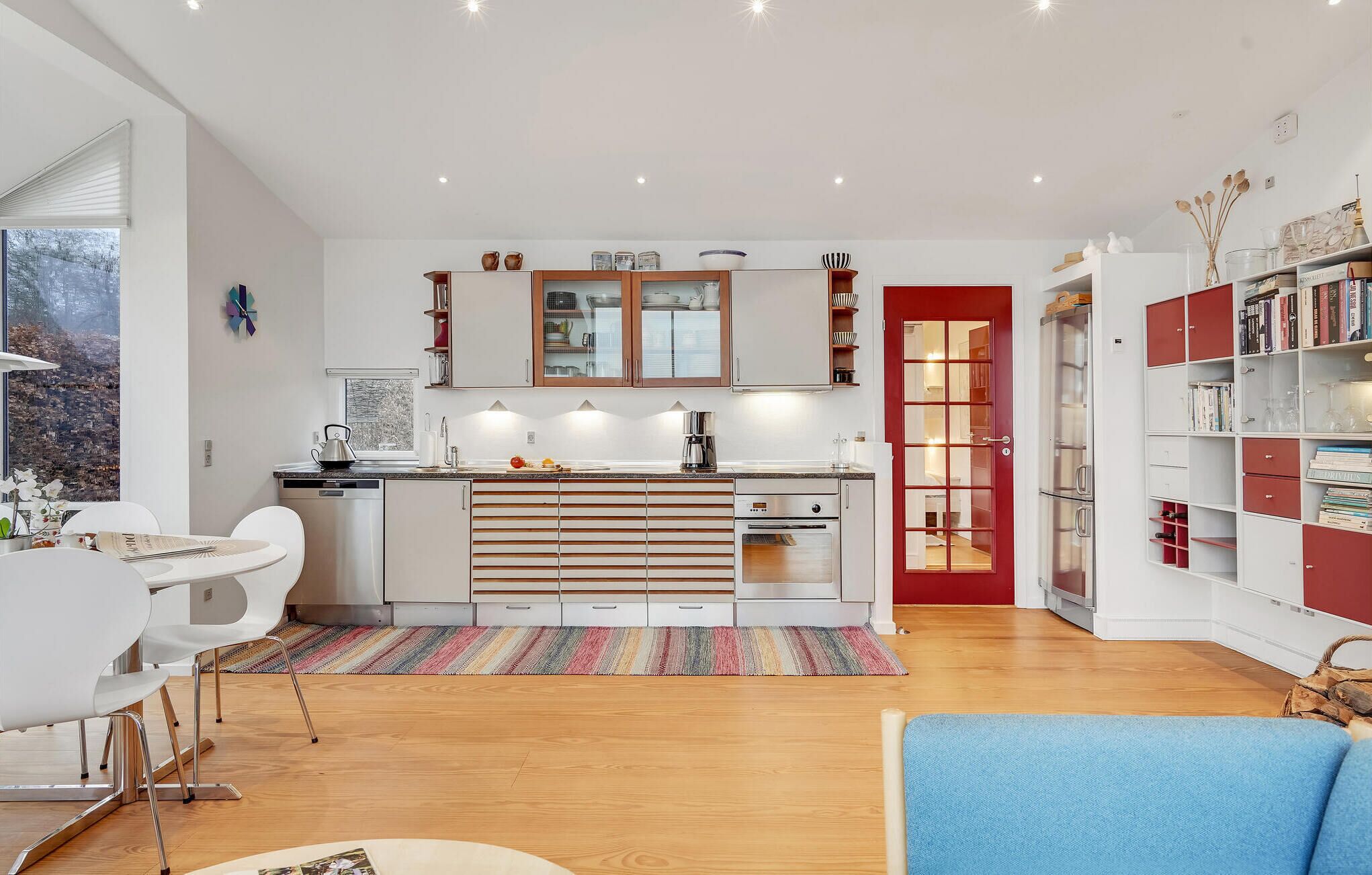 Une cuisine moderne avec des meubles blancs, une porte rouge et un coin repas avec des chaises blanches.