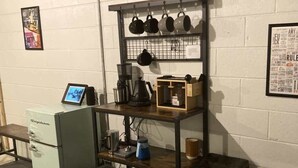 A kitchenette with a refrigerator, coffee maker, kettle, and shelves with mugs and a kettle.