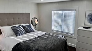 A bedroom with a bed, a bedside lamp, a window with blinds, and a dresser.
