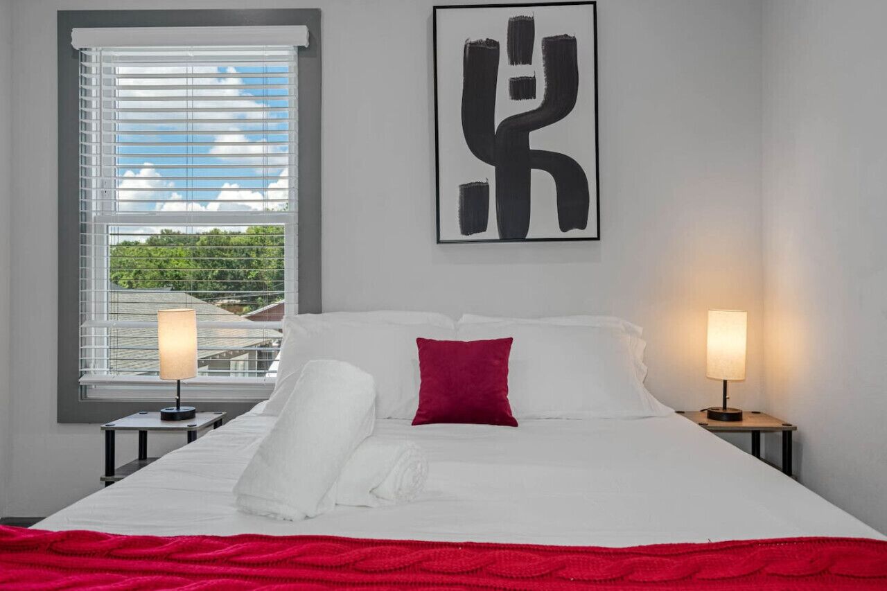 A hotel room with a bed, two bedside lamps, a red pillow, a white blanket, and a window with blinds.
