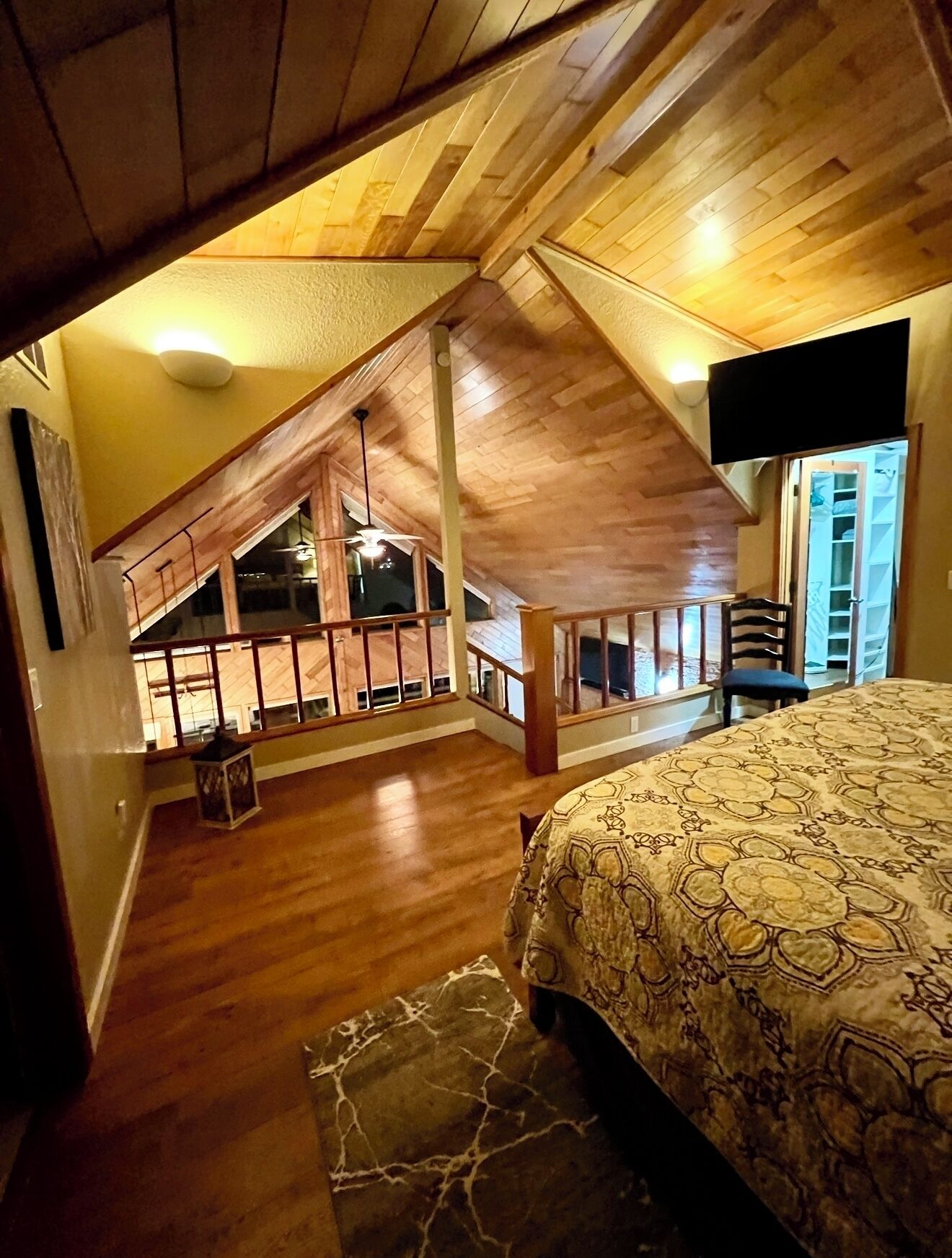 Un dormitorio con techo inclinado, piso de madera, cama con colcha estampada, una mesita y una silla.