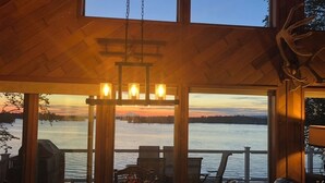 Una cabaña acogedora con interior de madera, un comedor con mesa y sillas, y un amplio ventanal con vistas a un lago al atardecer.