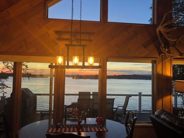 Una cabaña acogedora con interior de madera, un comedor con mesa y sillas, y un amplio ventanal con vistas a un lago al atardecer.