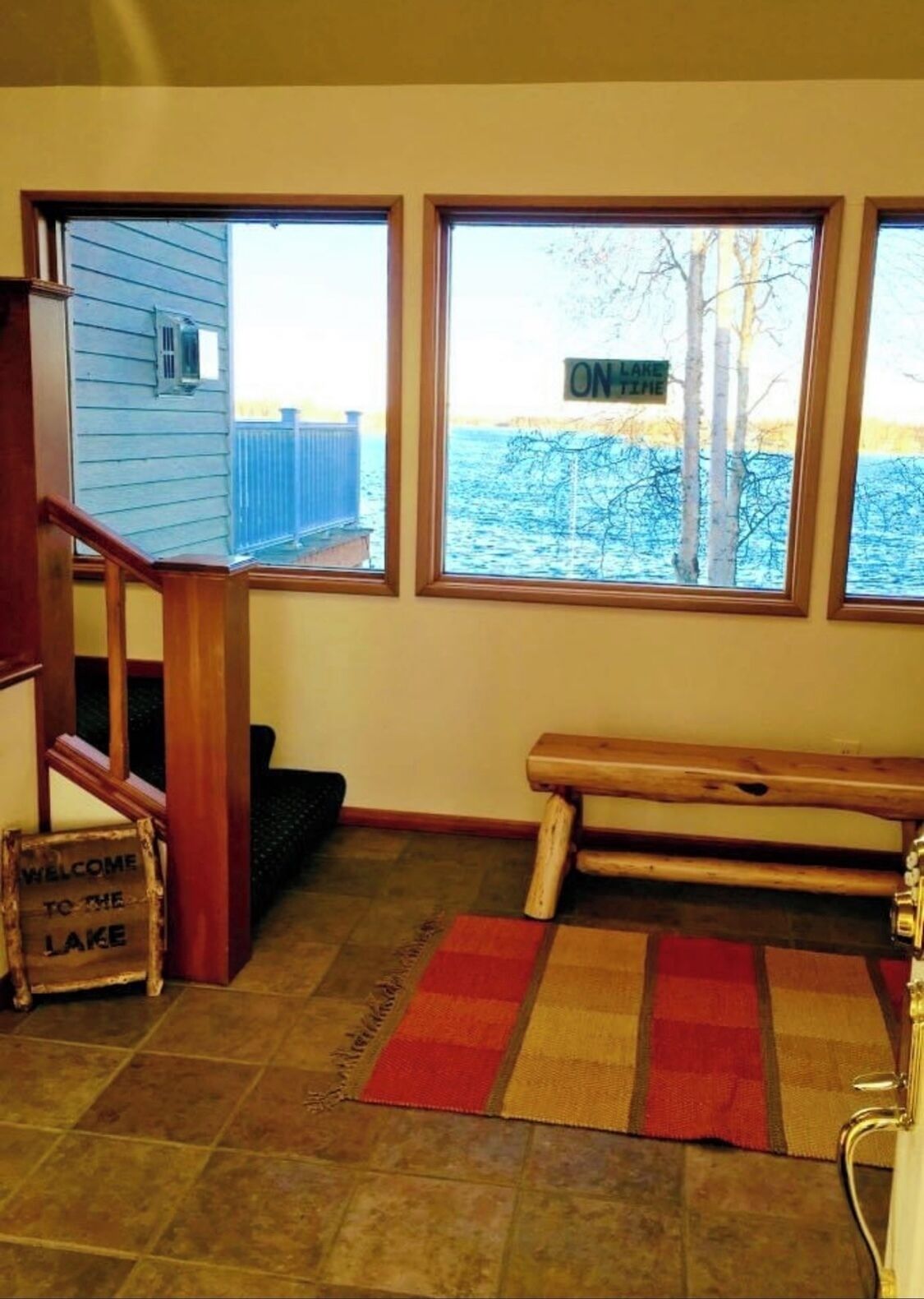 A room with a wooden bench, a welcome sign to the lake, and a view of the lake through large windows.