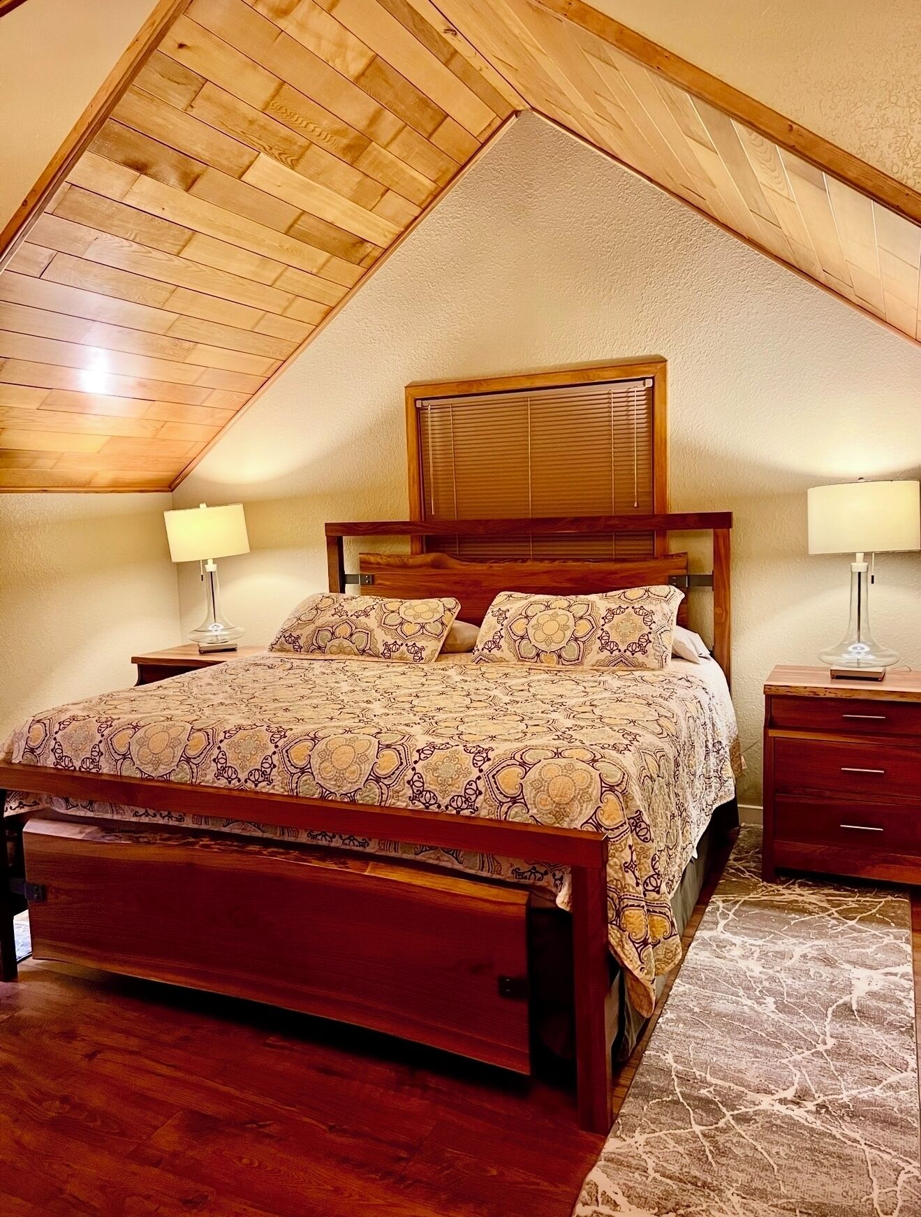 A bedroom with a sloped ceiling, wooden bed frame, patterned bedspread, two bedside tables with lamps, and a window with blinds.