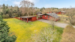 Une cabane rouge avec un porche couvert et un petit abri de jardin, entourée d’une cour verdoyante et d’arbres.