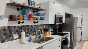 Una cocina moderna con armarios blancos, lavavajillas de acero inoxidable, microondas y horno empotrado. Hay tazas de colores en un estante, una tabla de cortar de madera y un tapete azul con conchas marinas en el suelo.