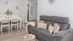 A modern living room with a grey sofa, white chairs, a small round table with books and vases, and a dining area with a white table and chairs. - Casa Moreno Cómpeta by Ruralidays (Málaga)