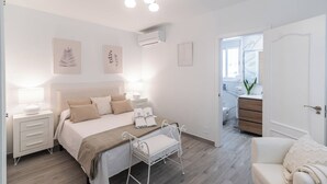 A modern bedroom with a bed, bedside tables, a nightstand, a chair, and a bathroom visible through an open door. - Casa Moreno Cómpeta by Ruralidays (Málaga)