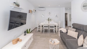 A modern living room with a flat-screen TV, a grey sofa, a dining table with chairs, and a small side table with books and a plant. - Casa Moreno Cómpeta by Ruralidays (Málaga)