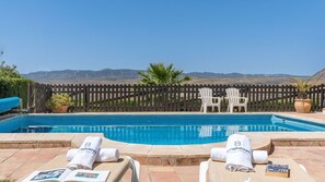 A swimming pool with a view of wind turbines in the background, surrounded by a wooden fence and white chairs. - El Collado de Lecrín El Padul by Ruralidays (Granada)
