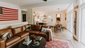 A spacious living room with a brown leather sofa, a coffee table with books and decorative items, a large American flag on the wall, and a kitchen in the background. - Antique Charm 8 Mins to Stratton 12 to Mt Snow (Stratton)